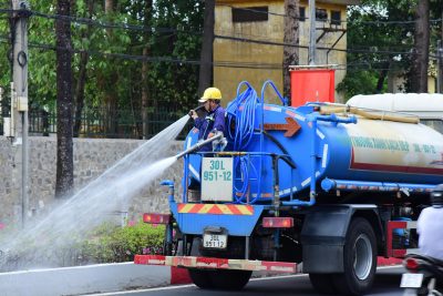 A worker operates a water truck in an urban setting, spraying water on the street. Un ouvrier fait fonctionner un camion-citerne d'eau en milieu urbain, pulvérisant de l'eau sur la rue.