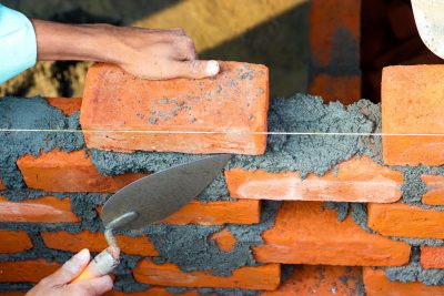 Hands laying bricks with cement in a construction setting, showcasing precise masonry work. Des mains posant des briques avec du ciment dans un cadre de construction, mettant en évidence un travail de maçonnerie précis.