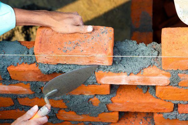 Hands laying bricks with cement in a construction setting, showcasing precise masonry work. Des mains posant des briques avec du ciment dans un cadre de construction, mettant en évidence un travail de maçonnerie précis.